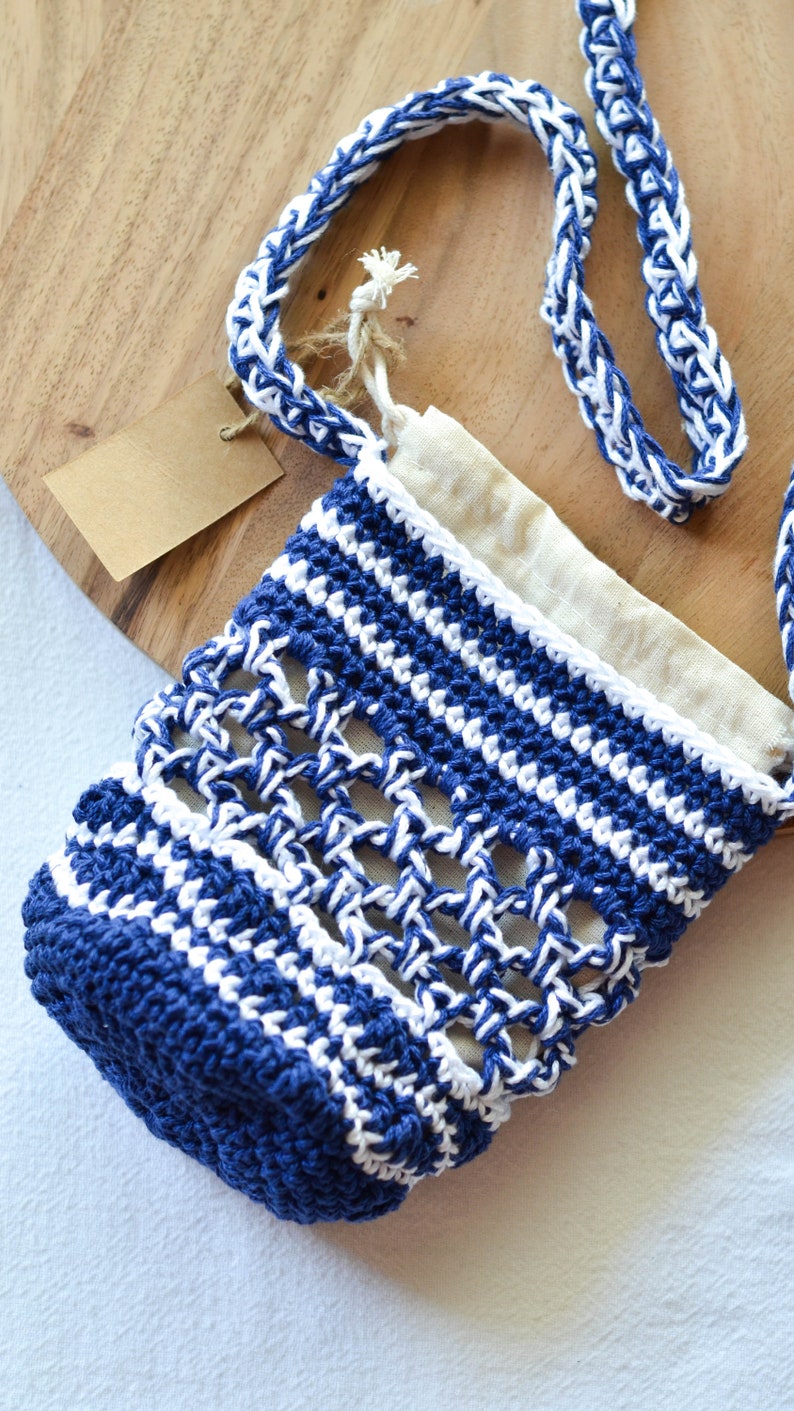 May include: A handmade blue and white crochet bag with a matching strap. The bag has an open weave design and a beige fabric lining. A small tag is attached to the bag.