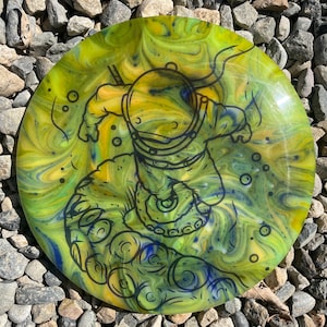 May include: A disc golf disc in shades of yellow, blue, and green with a swirling design. It has a black outline of an astronaut with tentacles. The disc is resting on a bed of small stones.