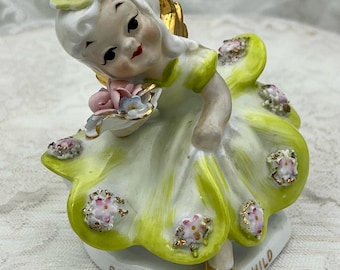 May include: A vintage porcelain figurine of a child in a yellow dress with gold accents, holding a basket of flowers. The figurine has a heart-shaped base with the text "SATURDAY'S CHILD". The child has blonde hair, rosy cheeks, and gold wings.