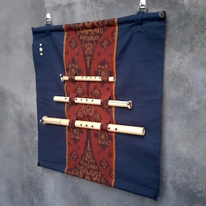 May include: A dark blue fabric wall hanging featuring a central vertical panel with a rust-coloured ikat pattern. Three light-coloured wooden flutes are held horizontally in fabric loops across the centre. The piece is suspended by two metal rings.