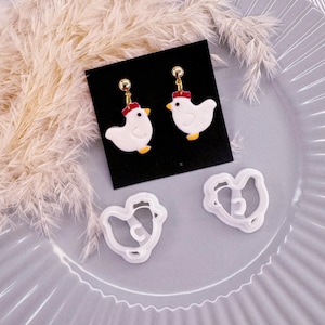 May include: A pair of white chicken earrings with red combs and gold accents, displayed on a black card. Two white plastic chicken cookie cutters are also shown on a grey surface.