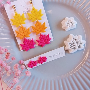 Puede incluir: Pendientes colgantes con diseños de hojas de arce amarillas, naranjas y rosas. Los pendientes se muestran en una tarjeta blanca con un gráfico de corona. También se muestran dos cortadores con forma de hoja y una tarjeta con "THANK YOU".