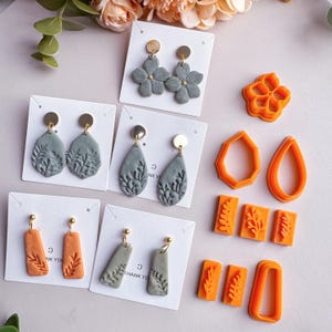 Puede incluir: Una variedad de pendientes hechos a mano en varias formas y colores, incluyendo diseños florales y de hojas grises, y formas rectangulares y geométricas naranjas. Se ven postes de pendientes dorados. También se incluyen cortadores de arcilla naranja.