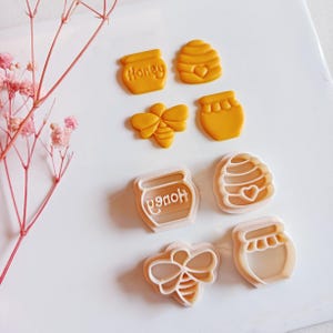 May include: A collection of yellow cookie cutters in the shape of honey pots, a bee, and a beehive. One honey pot has the word "Honey" on it. The other cookie cutters are white.