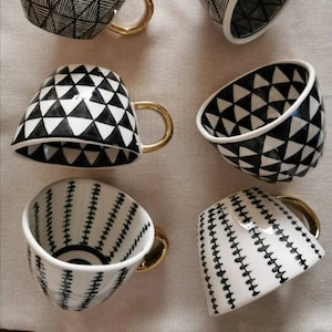 May include: Six black and white ceramic mugs with gold handles. The mugs have geometric patterns of triangles and lines.