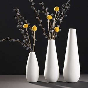 Könnte beinhalten: Drei weiße Keramikvasen in verschiedenen Höhen, mit einer schmalen, länglichen Form. Die Vasen sind in einer Reihe vom kleinsten zum größten angeordnet. Die größte Vase ist mit getrockneten gelben Blumen gefüllt.
