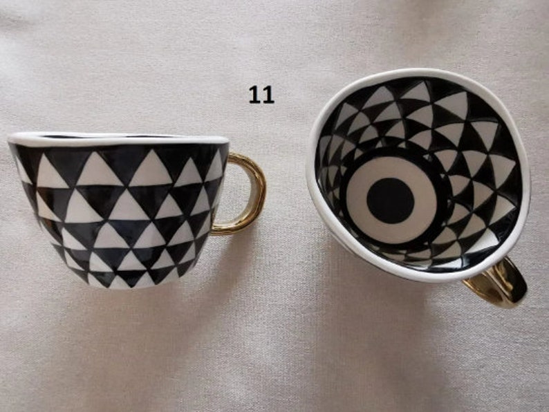 May include: A black and white ceramic coffee mug with a geometric pattern and a gold handle. The inside of the mug has a black and white eye design.