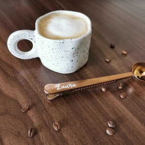 May include: A white ceramic coffee mug with black speckles and a gold-toned coffee scoop with a clip that says "Laura". The scoop is on a wooden surface with coffee beans scattered around.