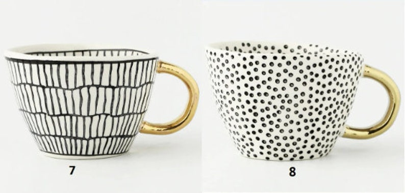 May include: Two white ceramic mugs with gold handles. The mug on the left has a black and white geometric pattern. The mug on the right has a black and white polka dot pattern.