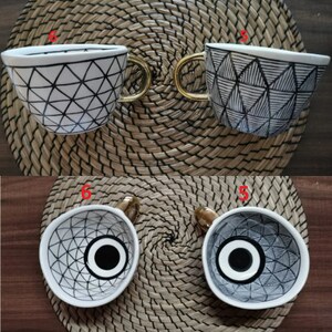 May include: Two white ceramic mugs with black geometric designs. The mugs have gold handles and are sitting on a woven mat.