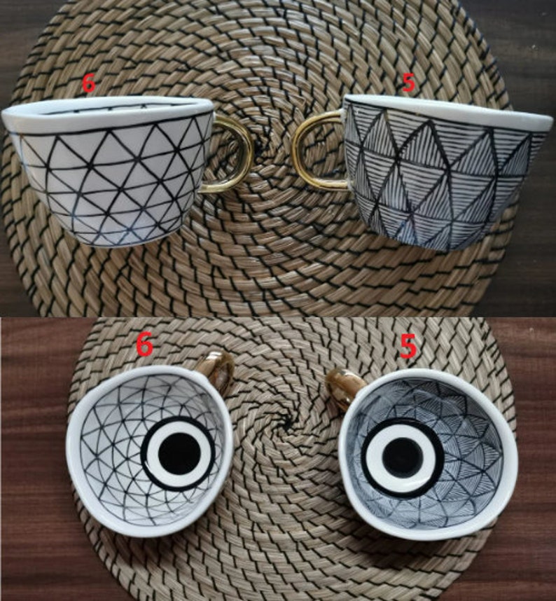 May include: Two white ceramic mugs with black geometric designs. The mugs have gold handles and are sitting on a woven mat.