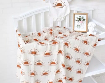 May include: A white muslin swaddle blanket with a repeating pattern of orange sunrises. The blanket is folded and draped over a white changing table.
