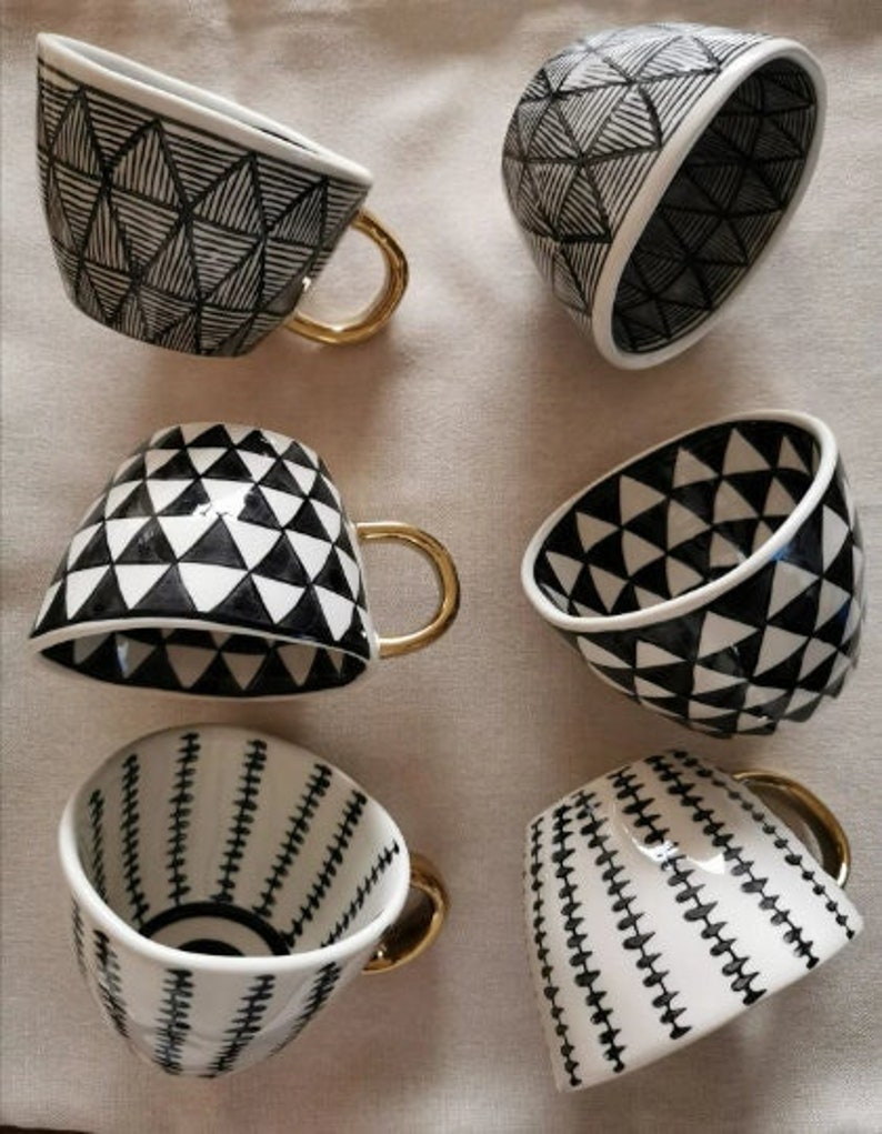 May include: Six black and white ceramic mugs with gold handles. The mugs have geometric patterns of triangles and lines.