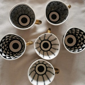 May include: Set of six white ceramic mugs with black and gold geometric designs. Each mug features a large black eye with a white center.