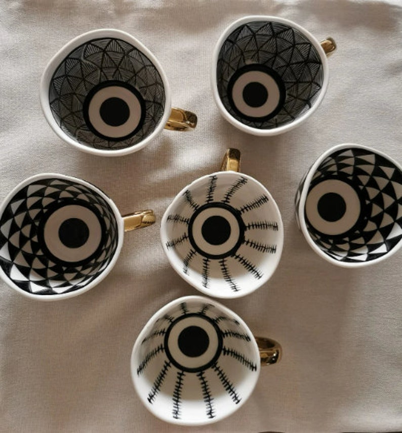 May include: Set of six white ceramic mugs with black and gold geometric designs. Each mug features a large black eye with a white center.