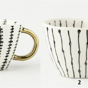 May include: Two white ceramic mugs with gold handles. The mugs have black and white geometric patterns. The mug on the left has a pattern of small black triangles. The mug on the right has a pattern of thin black lines with small black dots.