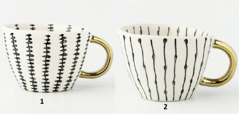 May include: Two white ceramic mugs with gold handles. The mugs have black and white geometric patterns. The mug on the left has a pattern of small black triangles. The mug on the right has a pattern of thin black lines with small black dots.