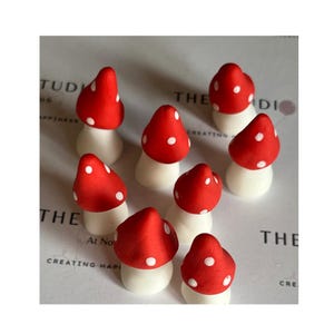 May include: A group of small mushroom ornaments. Each mushroom features a red cap with white spots and a white stem. The mushrooms are displayed on a light-coloured surface.