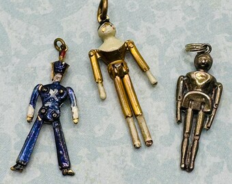 May include: Three vintage articulated doll charms. One is a blue and white soldier, one is a gold and white doll, and one is a silver doll.