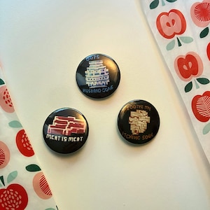 Peut inclure: Trois badges ronds à épingle sur fond noir. L'un présente une structure pixelisée bleue avec le texte OOPS HUSBAND GONE. Un autre montre de la viande pixelisée rouge avec le texte MEAT IS MEAT. Le troisième affiche un objet pixelisé beige avec le texte SOOTHE MY ITCHING SOUL.