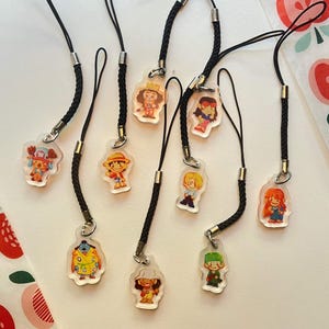 May include: A collection of nine clear acrylic charms featuring colourful cartoon character illustrations. Each charm is attached to a black braided cord with a silver metal connector, showcasing various figures with distinct outfits and hair colours.