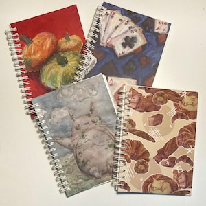 May include: Four different spiral bound notebooks with various designs. One notebook has a red cover with a painting of pumpkins. Another notebook has a blue cover with a painting of playing cards. A third notebook has a gray cover with a painting of a cartoon character. The fourth notebook has a beige cover with a painting of cats.