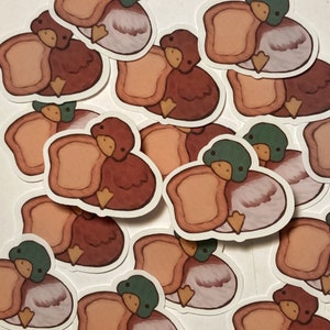 Puede incluir: Un conjunto de pegatinas con patos y pan de dibujos animados. Las pegatinas son de varios colores, incluyendo marrón, blanco y rosa. Los patos están representados con los ojos cerrados y expresiones sonrientes. El pan está representado como una hogaza con una corteza crujiente.