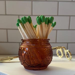 Puede incluir: Un pequeño tarro de vidrio color ámbar lleno de cerillas con puntas verdes. El tarro tiene un patrón decorativo en relieve. Un sujetador de cerillas dorado en forma de tijera descansa cerca. El tarro y las cerillas están sobre una pila de libros blancos.