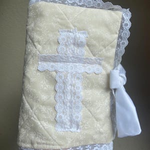 May include: Cream-colored quilted book cover with a floral pattern and white lace trim. A white lace cross is centered on the front. A white satin ribbon is tied on the side.