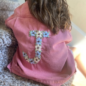 May include: A pink denim jacket with a floral embroidered "J" on the back. The embroidery features light blue, white, and cream-coloured flowers. The jacket is a solid pink colour and appears to be a child's size.