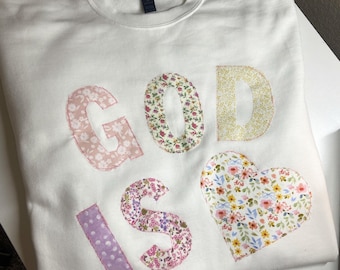 May include: White sweatshirt with the words "GOD IS" and a heart appliqued on the front. The letters and heart are made of floral patterned fabric in shades of pink, yellow, and purple.