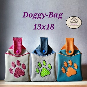 Op de afbeelding: Drie hondenkoekjeszakjes in roze, groen en oranje, elk met een pootafdrukontwerp. De zakjes zijn rechthoekig met een flap en een knoopsluiting. De tekst "Doggy-Bag 13x18" staat boven de zakjes.