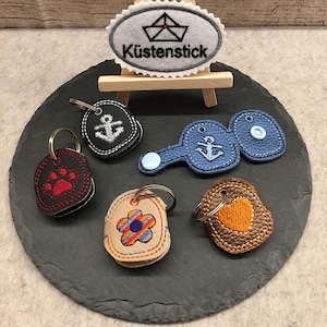 May include: Five different keychains with various embroidered designs. The keychains are made of leather and feature a paw print, a flower, a heart, and two anchors. The keychains are displayed on a black slate surface with a small wooden easel holding a white oval patch with the text "Küstenstick" and a small sailboat design.