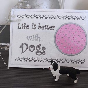 May include: White canvas sign with the text "Life is better with Dogs" in gray embroidery. A pink circle with a pattern of small hearts is in the center of the sign. A black and white border collie figurine is in the foreground.