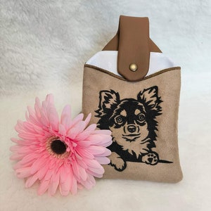 May include: A brown suede pouch with a white lining and a brown leather strap. The pouch features an embroidered image of a black chihuahua peeking out from the top.