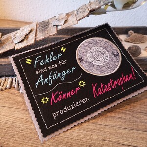 Op de afbeelding: Een zwarte vilt onderzetter met een witte gestikte rand en de tekst "Fehler sind was für Anfänger Können Katastrophen!" in kleurrijke borduurwerk.
