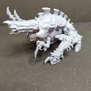 May include: A gray, detailed miniature of a creature with a segmented body, multiple limbs, and a large head with horns and a prominent jaw. The model has a textured surface and is posed on a dark surface.