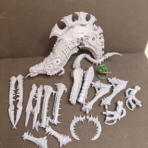Peut inclure: Ensemble de pièces de modèle en plastique gris clair non assemblées, comprenant une grande tête de créature détaillée et divers appendices épineux. Une petite figurine verte est également présente, suggérant un thème de jeu de guerre miniature.