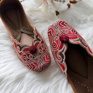 May include: A pair of red and gold embroidered flats with a brown leather sole. The flats feature a decorative pattern and a small red pom-pom. The shoes are displayed on a white, fluffy surface.