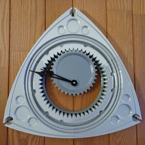 May include: A gray, triangular clock with a gear-shaped face and black hands. The clock is designed to look like a Mazda rotary engine.