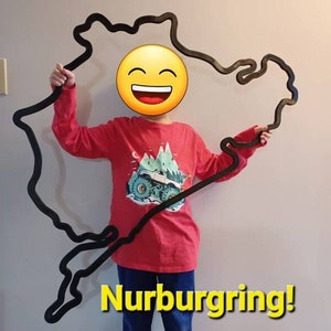 May include: A child holds up a large black outline of the Nurburgring race track. The track is made of wood and is about the size of the child. The child is wearing a red long-sleeved shirt with a mountain and truck graphic. The text "Nurburgring!" is written in yellow letters at the bottom of the image.