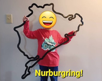 May include: A child holds up a large black outline of the Nurburgring race track. The track is made of wood and is about the size of the child. The child is wearing a red long-sleeved shirt with a mountain and truck graphic. The text "Nurburgring!" is written in yellow letters at the bottom of the image.