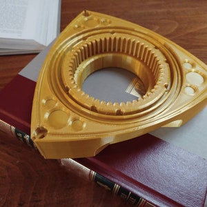 May include: A gold-colored, three-pointed, rotary engine housing replica. The housing has a gear-like design and a central opening.