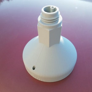 May include: White plastic 3D printed object with a hexagonal base and a round top with a small hole. The object has a threaded top.