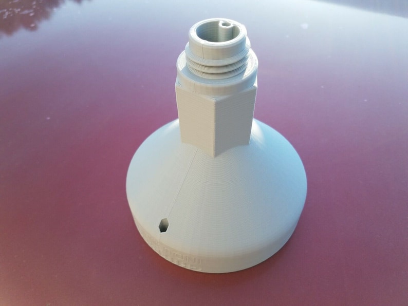 May include: White plastic 3D printed object with a hexagonal base and a round top with a small hole. The object has a threaded top.