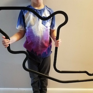 May include: A black metal outline of a race track, held by a person. The track is large and appears to be a wall hanging.