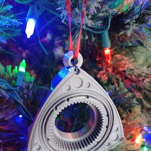 May include: A silver-colored, triangular-shaped Christmas ornament with a gear-like design in the center. The ornament is hanging from a red ribbon on a Christmas tree.
