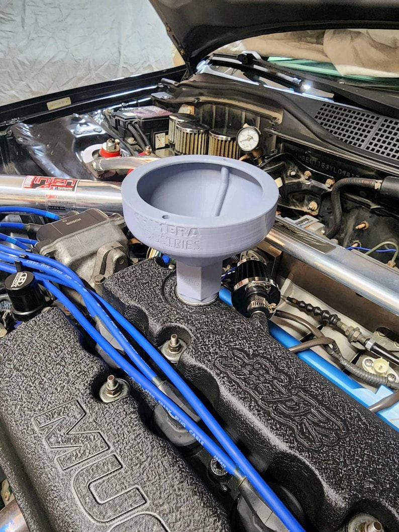May include: A grey plastic oil funnel with the text "TERA INDUSTRIES" printed on the side. The funnel is inserted into the opening of a car engine.