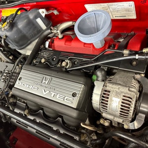 May include: Close-up of a red car engine with a black plastic engine cover that says "DOHC VTEC HONDA MOTOR CO.". The engine has a silver alternator and a grey funnel.