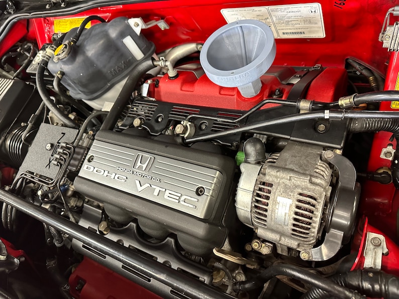 May include: Close-up of a red car engine with a black plastic engine cover that says "DOHC VTEC HONDA MOTOR CO.". The engine has a silver alternator and a grey funnel.